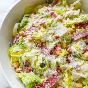 Copycat La Scala Chopped Salad mixed with lettuce, salami, chickpeas, and mozzarella, topped with cheese and black pepper in a white bowl.