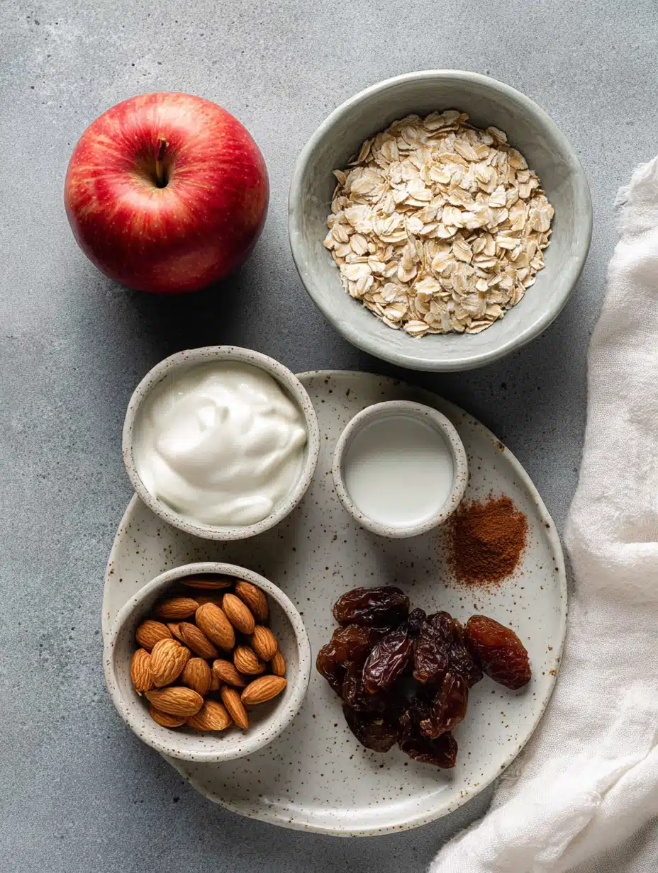 Flat lay of Bircher Muesli recipe ingredients including apple, oats, yogurt, almonds, and raisins on gray stone.