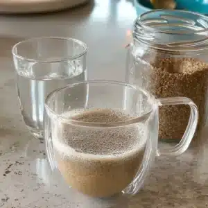Double-walled glass cup filled with Bariatric Seed Trick drink, cloudy and foamy on a marble counter with seed mix jar.