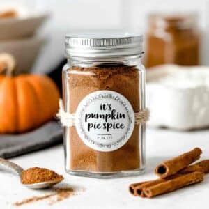 Jar of pumpkin pie spice with cinnamon sticks and spoon on white background