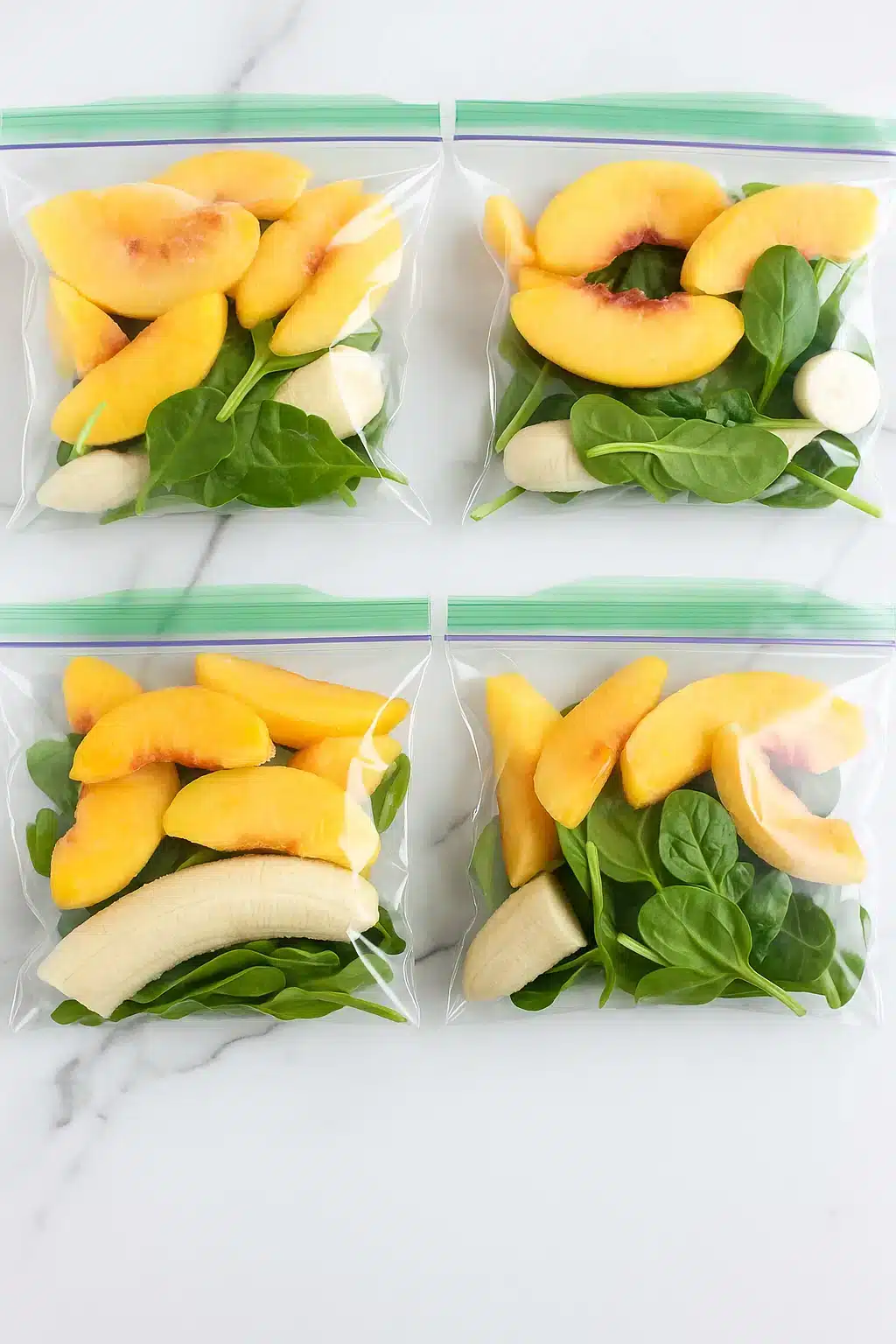 Four ginger peach smoothie freezer bags on a marble counter, each filled with peach slices, spinach, and banana.