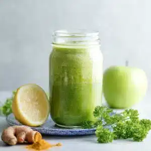 Green apple spinach smoothie in a mason jar on a blue patterned plate with lemon, green apple, ginger root, turmeric, and parsley.